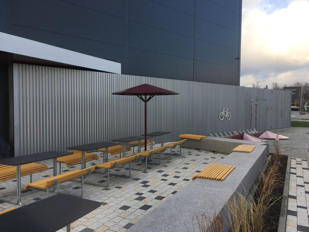 Nowoczesna przestrzeń publiczna z metalowym ogrodzeniem, stołami piknikowymi z drewnianymi ławkami, stojakiem na rowery i dekoracyjnym murkiem z roślinami, na tle budynku o ciemnej elewacji.