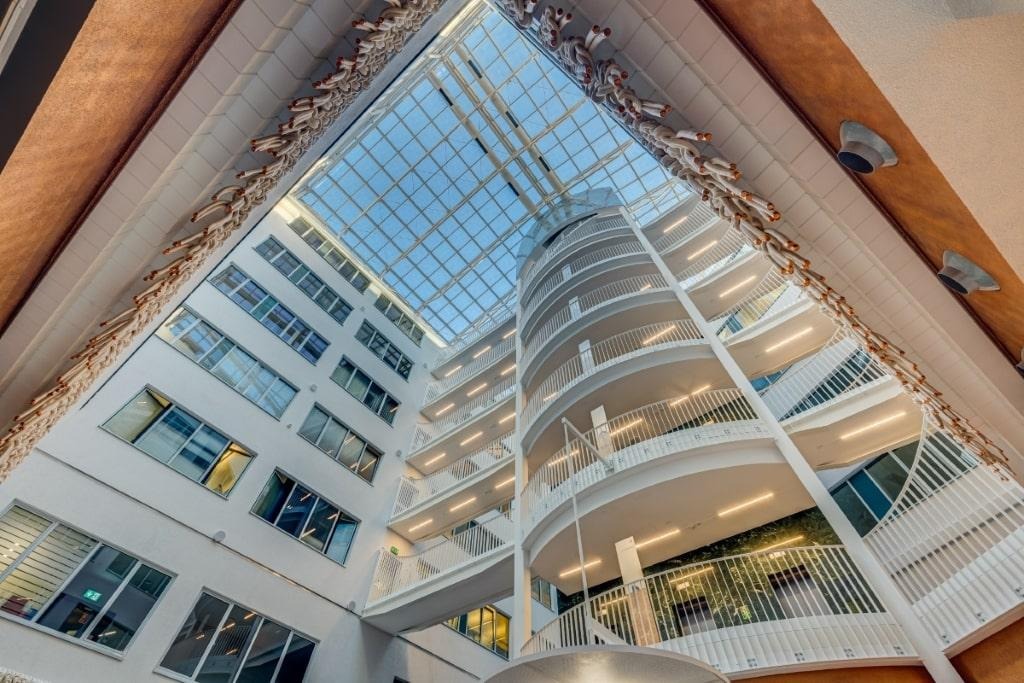 Wnętrze nowoczesnego budynku z przeszklonym dachem, spiralnymi schodami i białymi balustradami, widok z dołu na górę.