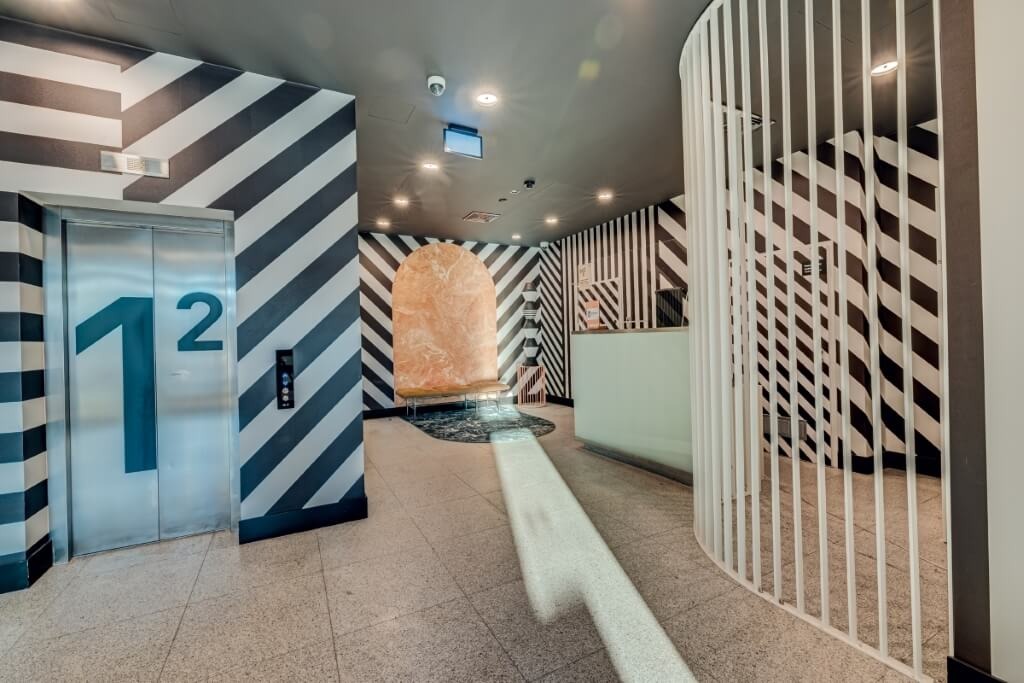 Nowoczesne wnętrze holu z metalową obudową windy oznaczoną numerami 1 i 2, białą, ażurową balustradą i dekoracyjnymi elementami architektonicznymi na ścianach w pasy.