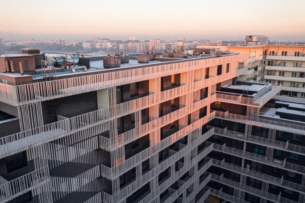 Wielopiętrowy budynek mieszkalny z balkonami zabezpieczonymi białymi, pionowymi barierkami, widok z góry w porannym świetle.
