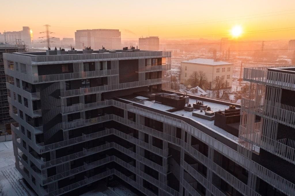 Nowoczesny budynek mieszkalny z balkonami zabezpieczonymi białymi, pionowymi barierkami, widziany z góry w porannym świetle, z miejskim krajobrazem w tle.