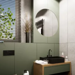 Nowoczesna łazienka w odcieniach zieleni i szarości z czarną armaturą i umywalką, okrągłe lustro z odbiciem grzejnika, minimalistyczny design.