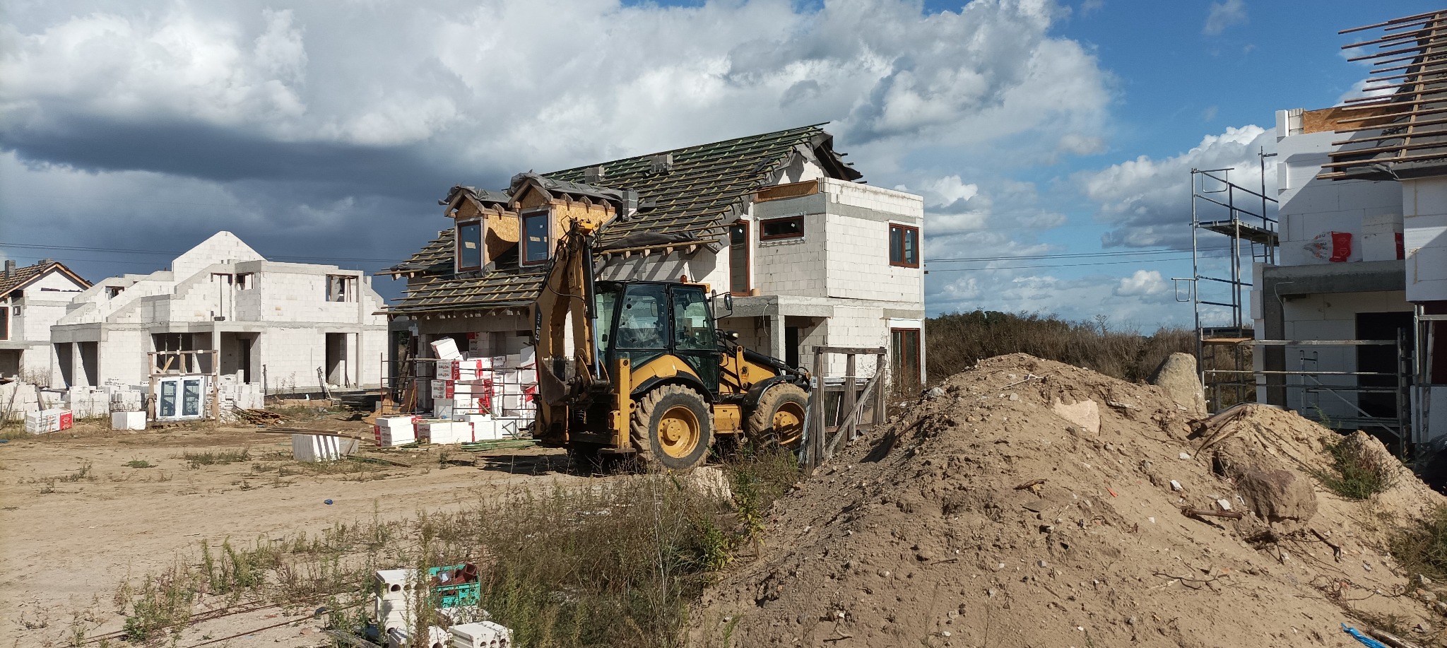 Budowa domu jednorodzinnego w stanie surowym otwartym, z widocznym żółtym koparko-ładowarką na pierwszym planie i stertą ziemi, na tle pochmurnego nieba.