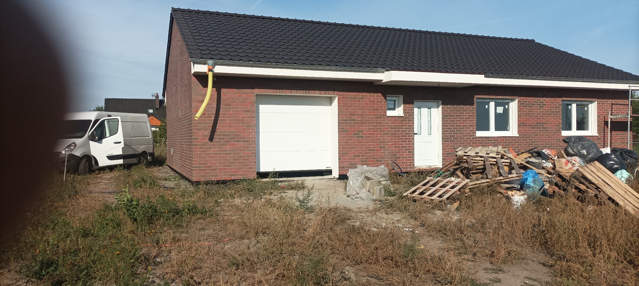 Nowo wybudowany dom z czerwonej cegły z białą bramą garażową i drzwiami wejściowymi, widoczne palety i worki z odpadami budowlanymi na trawniku, zaparkowany biały van w tle.