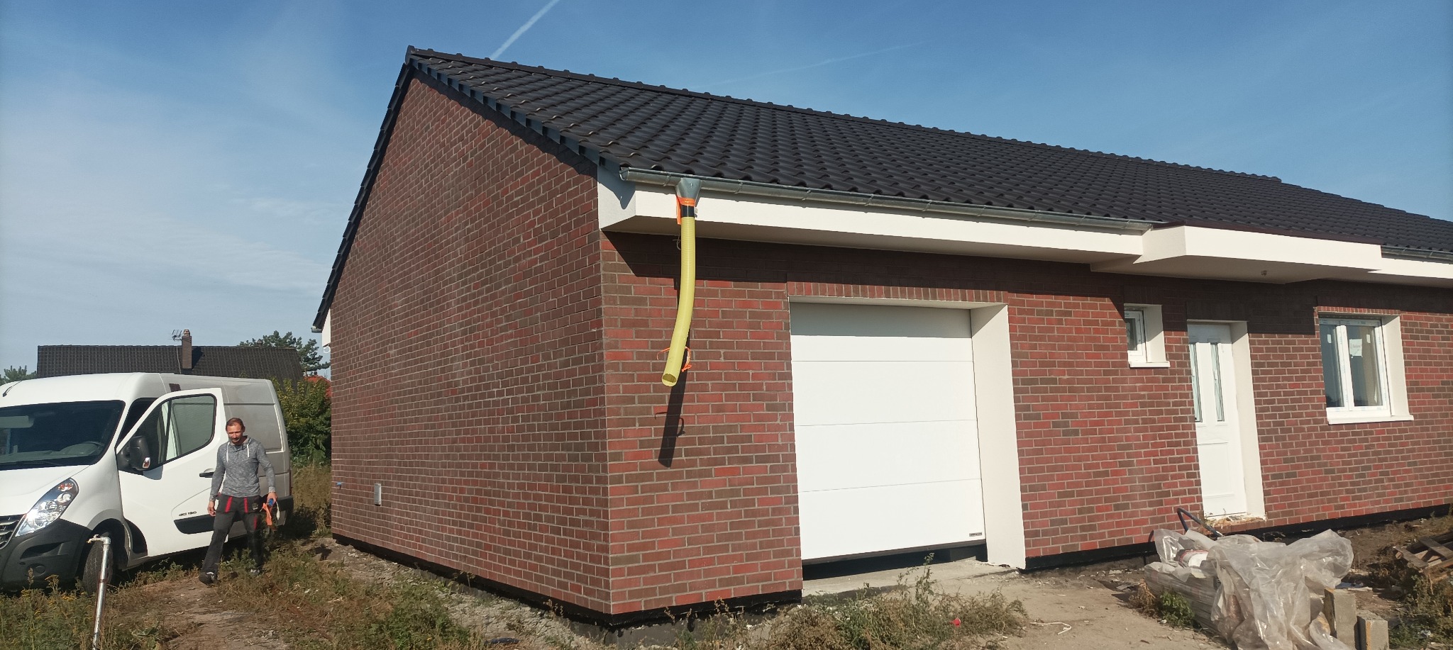 Nowo wybudowany budynek z czerwonej cegły z białą bramą garażową i drzwiami wejściowymi, widoczny pracownik z samochodem dostawczym w tle.