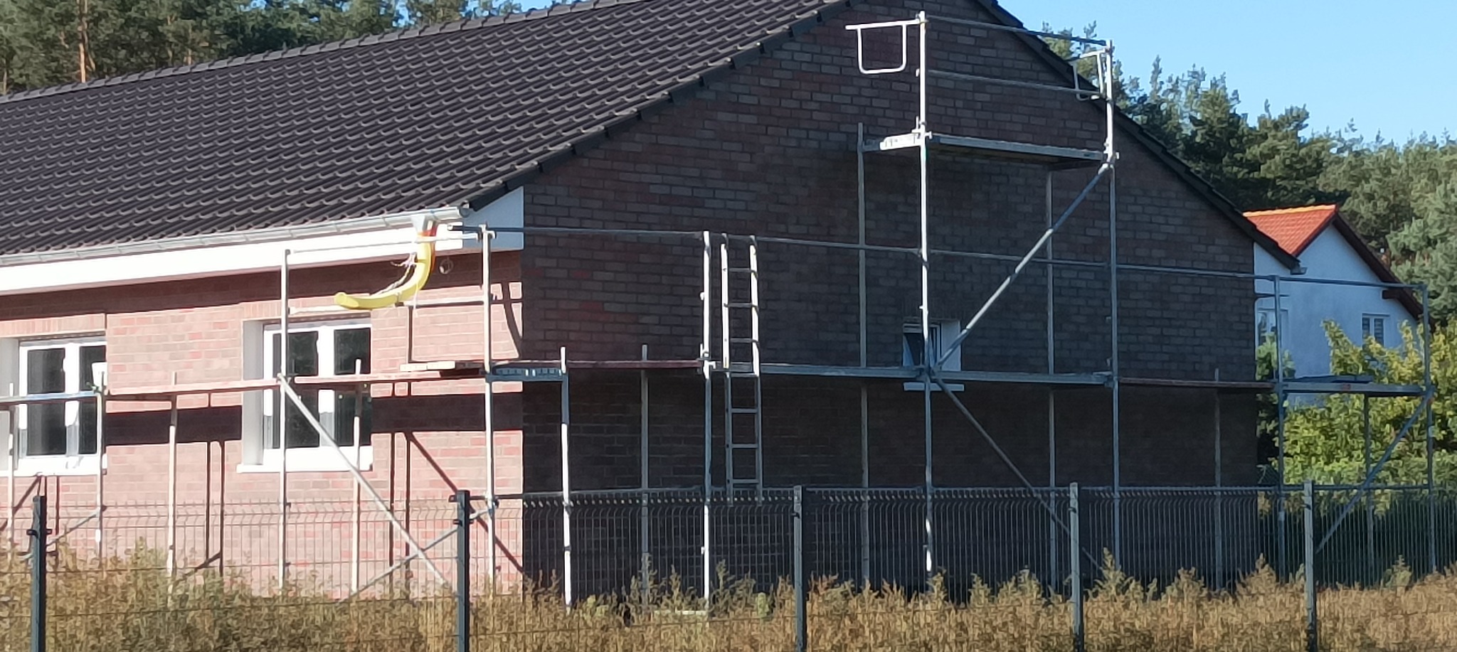 Dom jednorodzinny z cegły, częściowo otoczony rusztowaniem, z widocznymi elementami przygotowania do prac elewacyjnych, w tle drzewa i sąsiedni budynek.