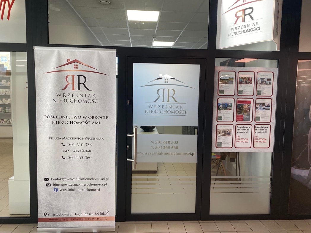Wejście do biura agencji nieruchomości Wrześniak, z widocznym roll-upem reklamowym, logo na szybie i ogłoszeniami w oknie.