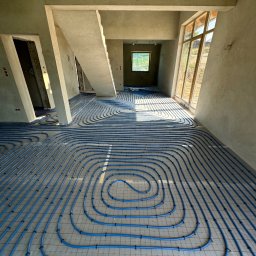 Expert Piotr Rzeszewicz - Niedokończona instalacja ogrzewania podłogowego z niebieskich rur na siatce, w surowym wnętrzu z wylewką i oknami.