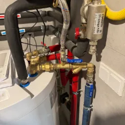 Instalacja grzewcza z widocznym filtrem Fernox TF1 Omega, rurami w izolacji czerwonej i niebieskiej, oraz elementami miedzianymi i zaworami. Widoczne również rury gazowe w żółtej osłonie karbowanej.