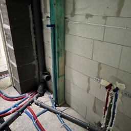 Expert Piotr Rzeszewicz - Wnętrze w trakcie budowy z widocznymi rurami instalacji wodnej (czerwone i niebieskie) oraz kanalizacyjnymi (szare) na podłodze i w ścianie z betonu komórkowego, przymocowanymi do ściany za pomocą...