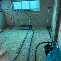 Expert Piotr Rzeszewicz - Surowy stan pomieszczenia w trakcie prac instalacyjnych: widoczne rury instalacji wodnej w niebieskiej karbowanej osłonie, poprowadzone po podłodze i ścianach z surowego betonu, puszki elektryczne...