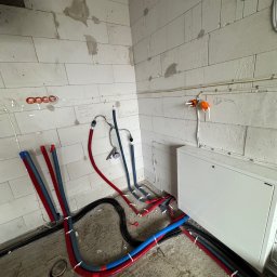 Expert Piotr Rzeszewicz - Surowe wnętrze w trakcie prac instalacyjnych: widoczne rury grzewcze i hydrauliczne w kolorach czerwonym i niebieskim, prowadzone po podłodze i ścianach z betonu komórkowego. Na ścianie widoczne...