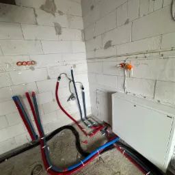 Surowe wnętrze w trakcie prac instalacyjnych: widoczne rury grzewcze i hydrauliczne w kolorach czerwonym i niebieskim, prowadzone po podłodze i ścianach z betonu komórkowego. Na ścianie widoczne...