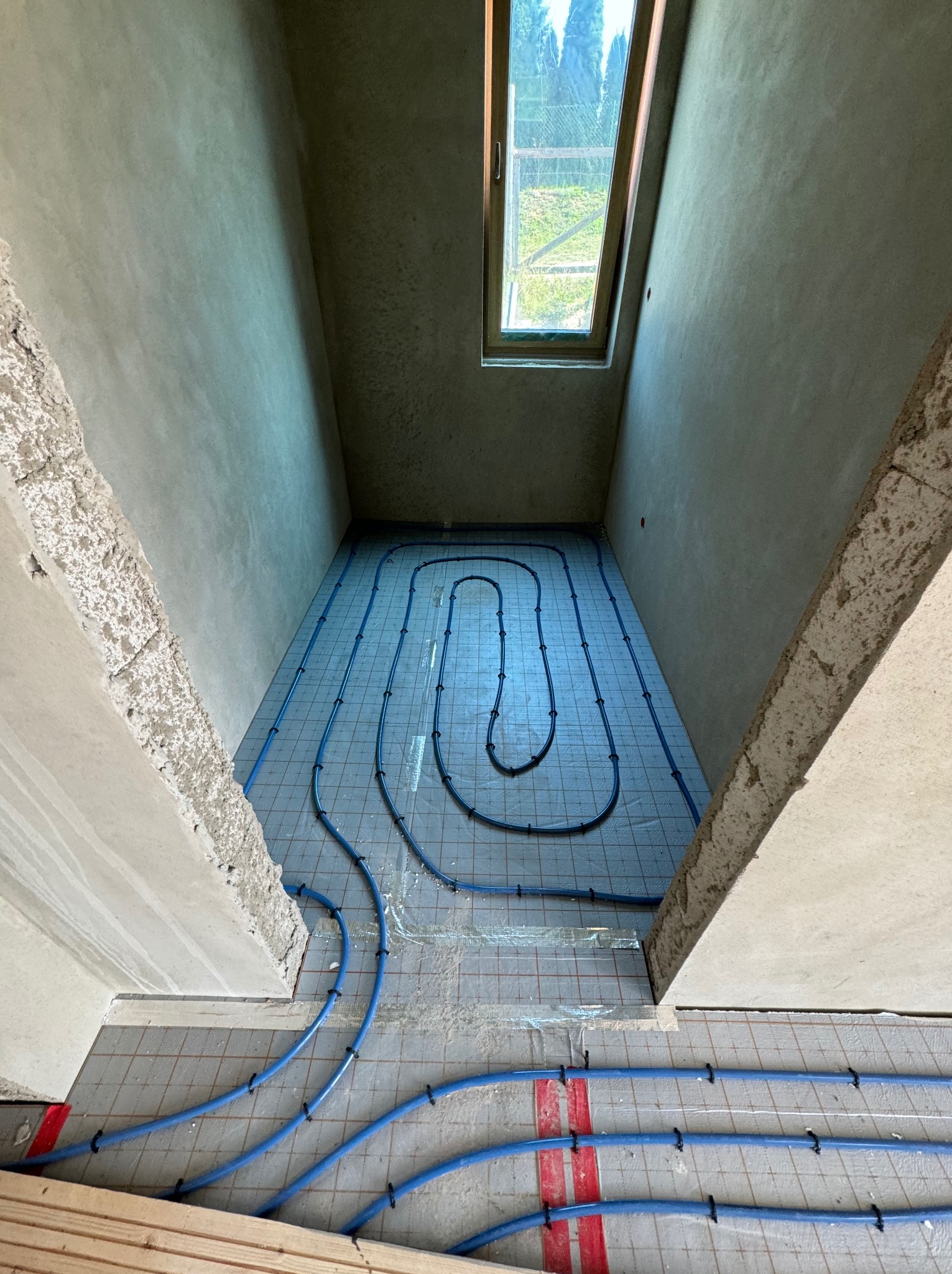 Widok z góry na nieotynkowane pomieszczenie z oknem dachowym, gdzie na podłodze ułożone są niebieskie rury ogrzewania podłogowego, przymocowane czarnymi uchwytami do siatki z czerwonymi paskami.
