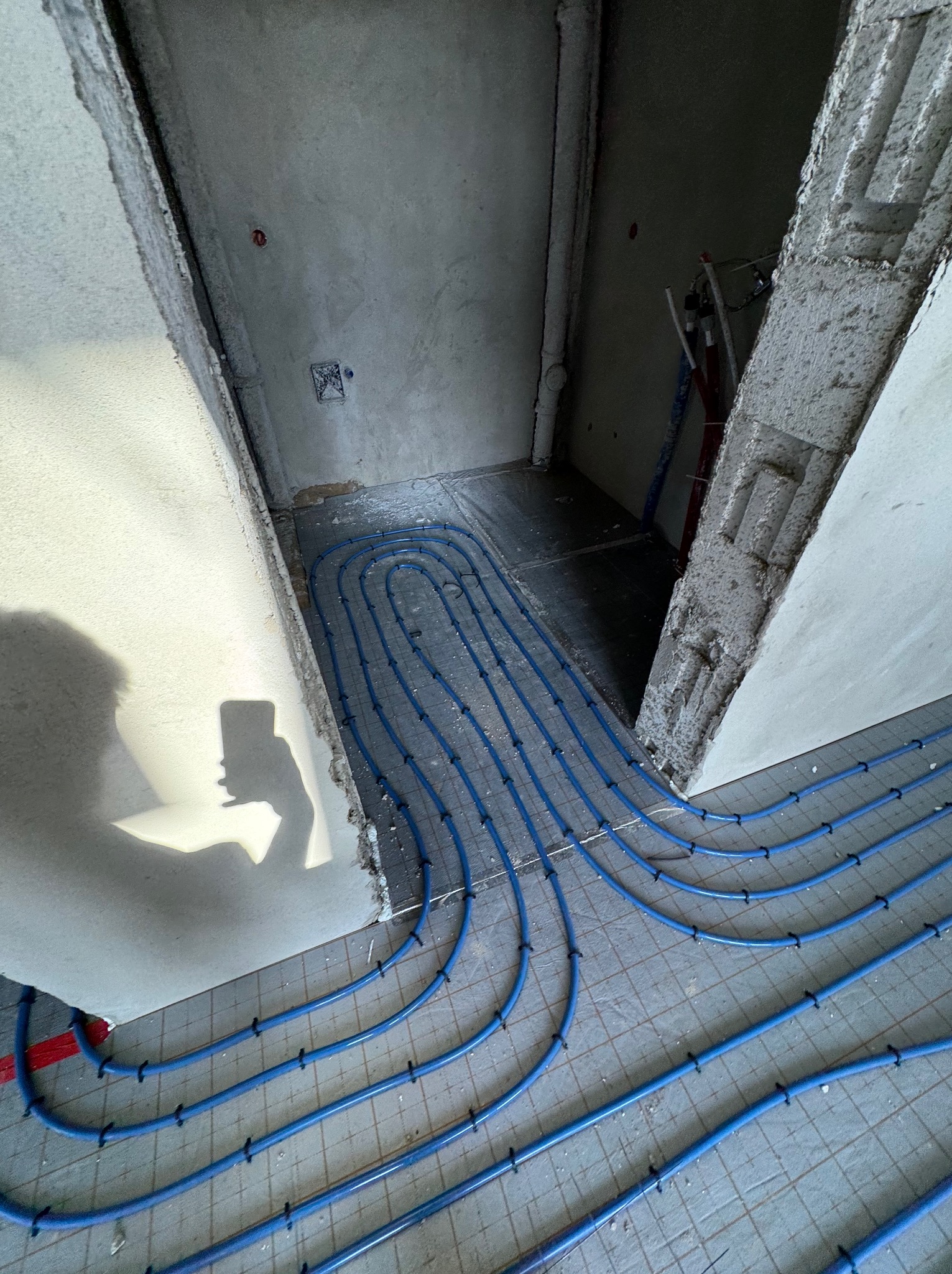 Niedokończona instalacja ogrzewania podłogowego z niebieskich rur na szarej podłodze, widoczny cień osoby robiącej zdjęcie telefonem, surowe ściany i niedokończone wykończenia.