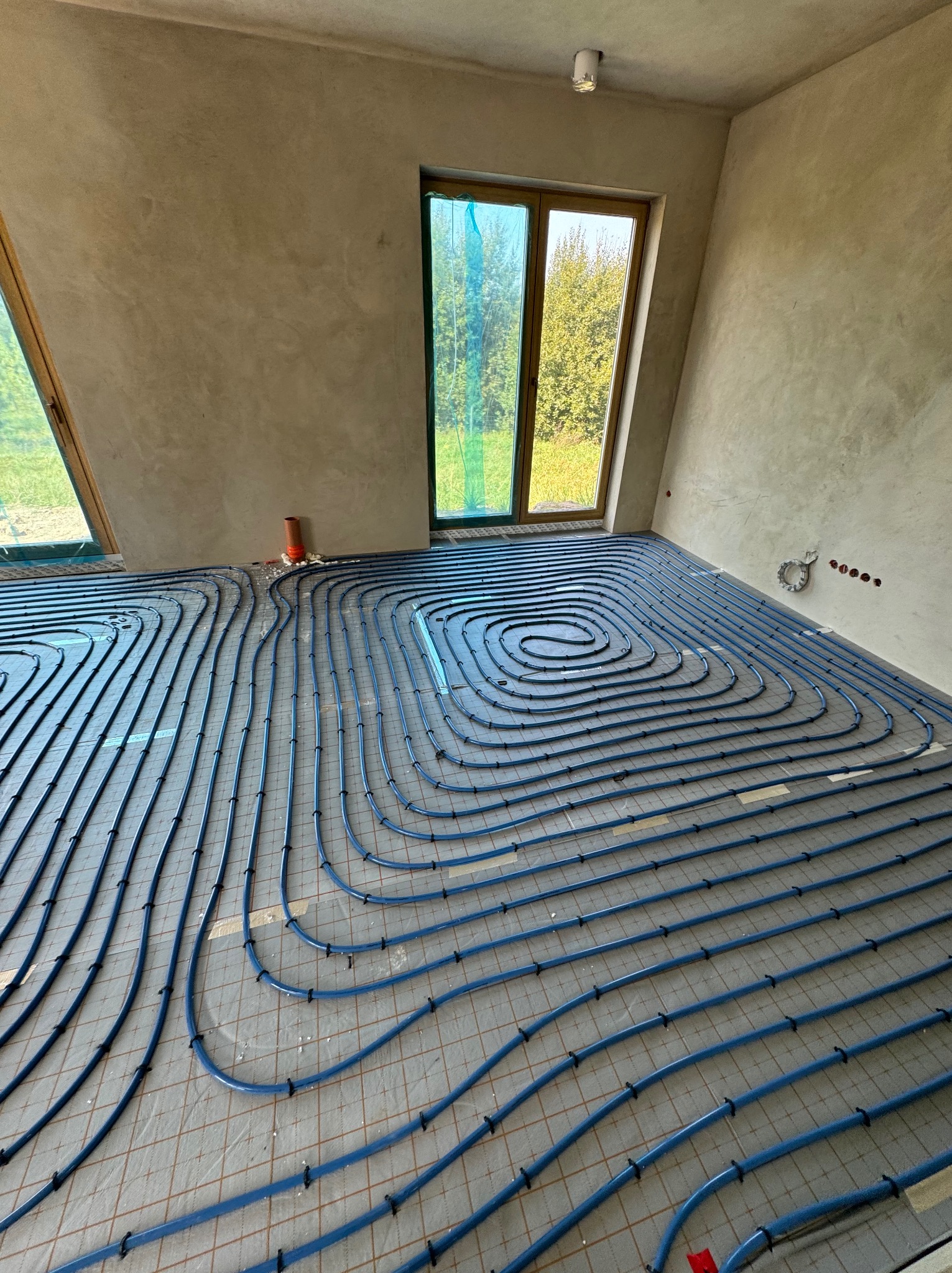 Niebieskie rury ogrzewania podłogowego ułożone spiralnie na siatce w surowym pomieszczeniu z oknami, widoczne elementy mocujące i przygotowane wyprowadzenia.