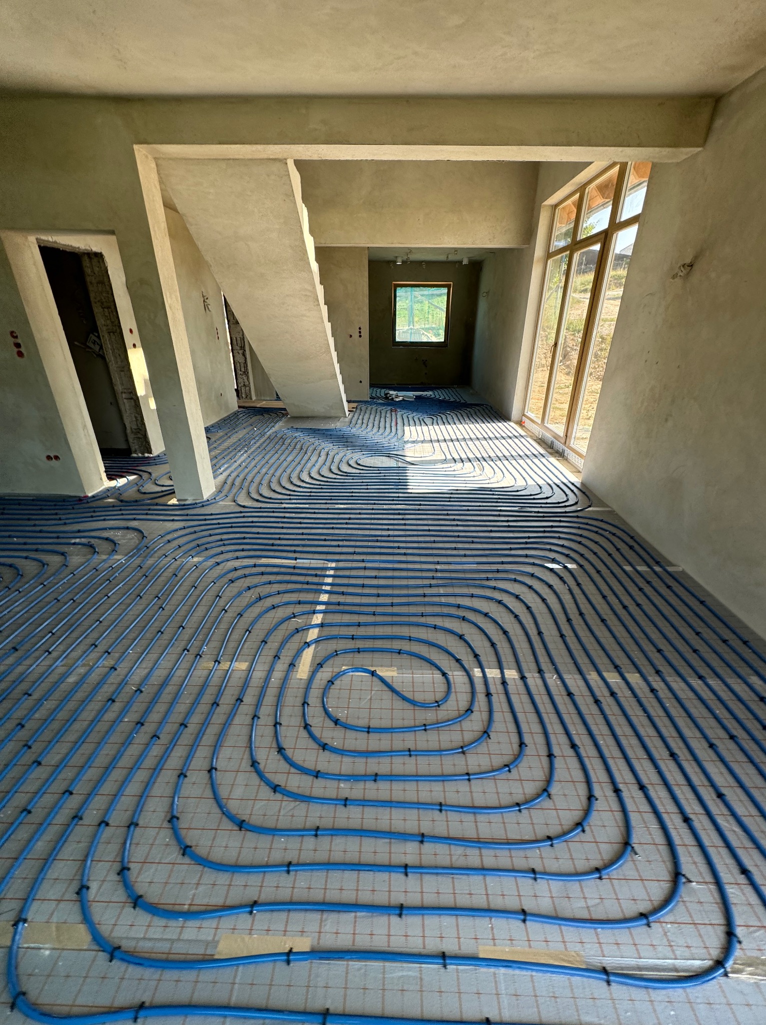 Niedokończona instalacja ogrzewania podłogowego z niebieskich rur na siatce, w surowym wnętrzu z wylewką i oknami.