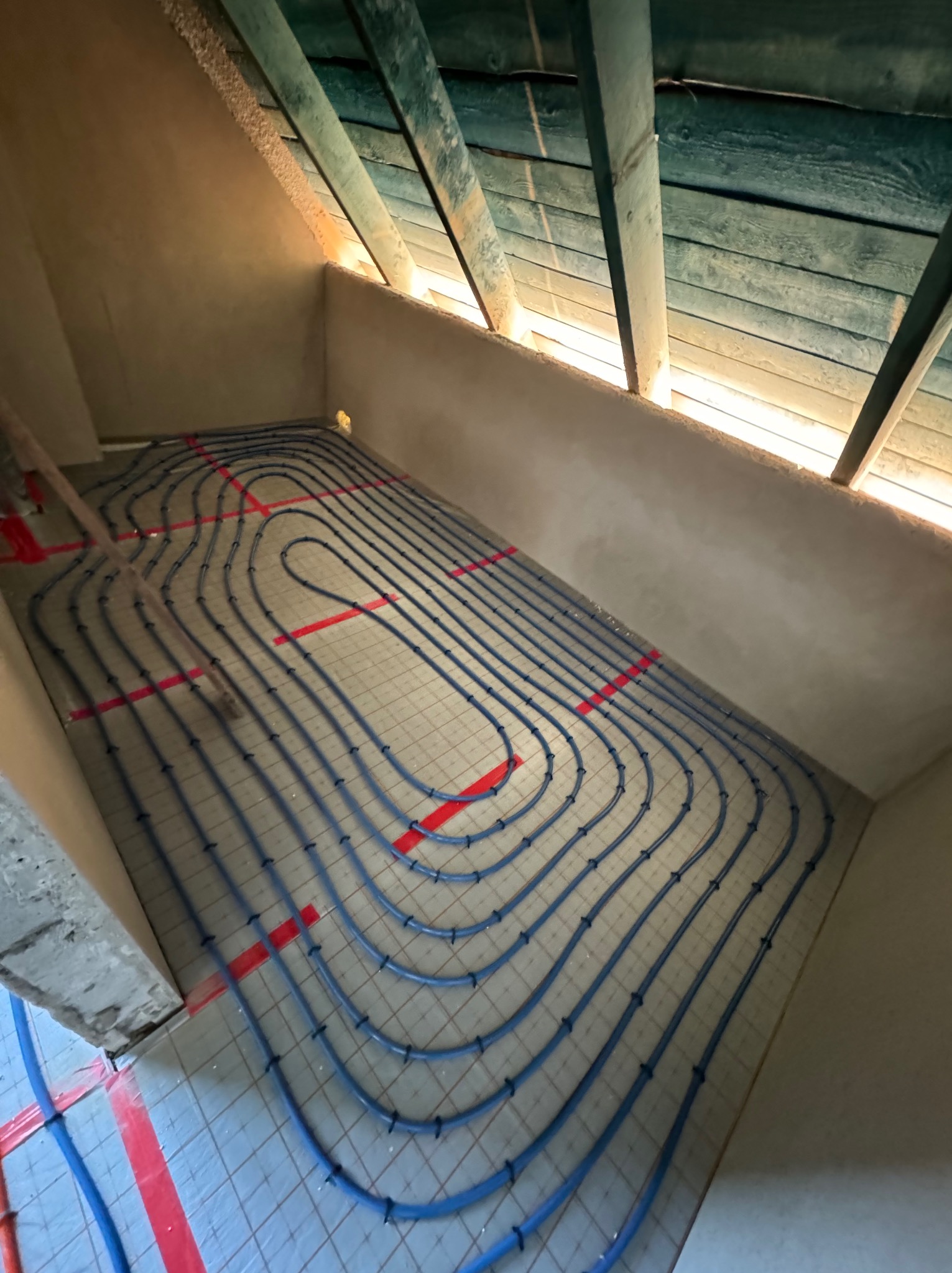 Niebieskie rury ogrzewania podłogowego ułożone na siatce w surowym pomieszczeniu na poddaszu w Gdańsku, widok z góry na instalację przed wylewką.