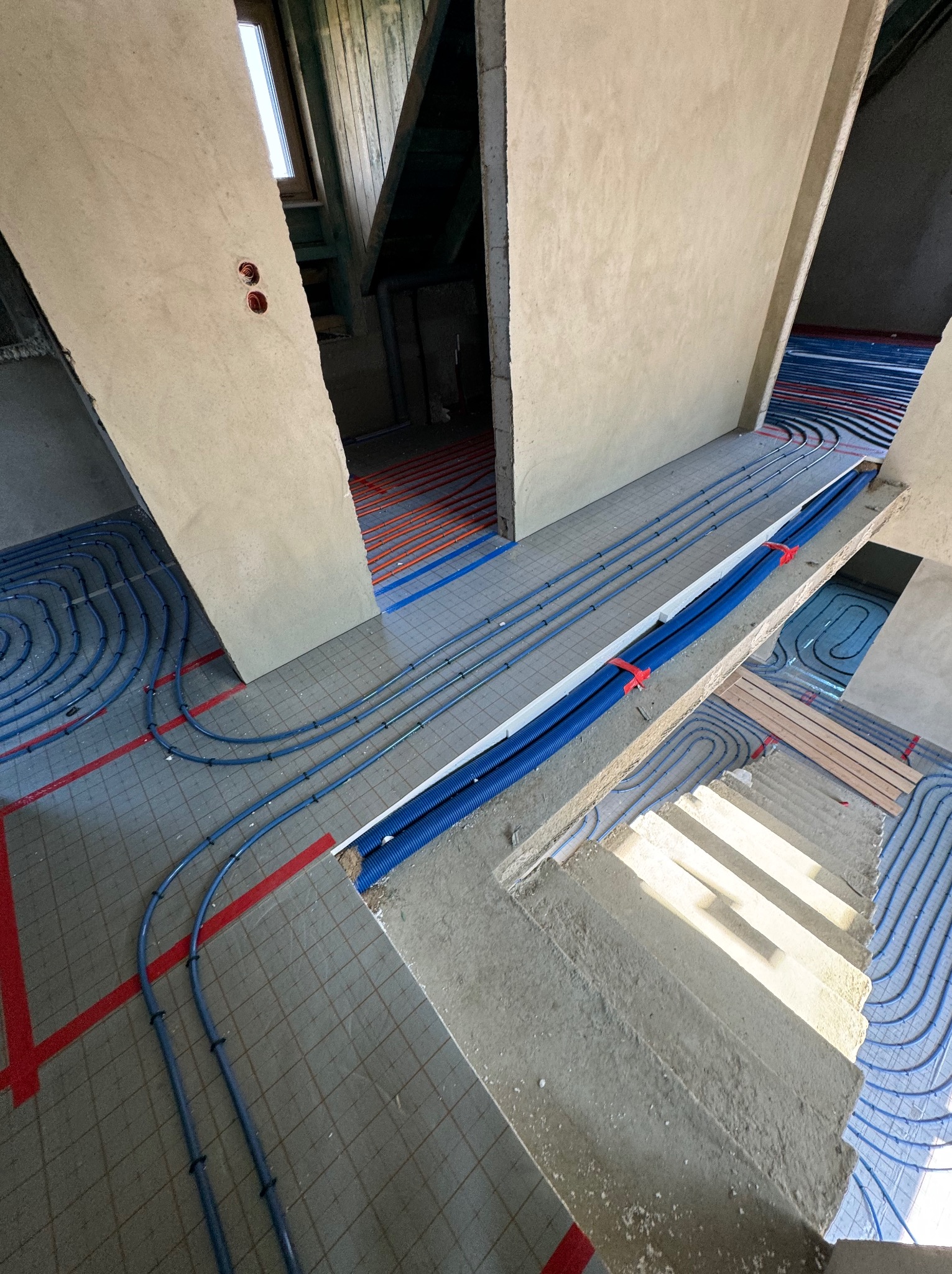 Widok z góry na instalację ogrzewania podłogowego w trakcie montażu w surowym wnętrzu, z widocznymi pętlami rur grzewczych, siatką montażową z czerwonymi liniami i izolacją termiczną,...