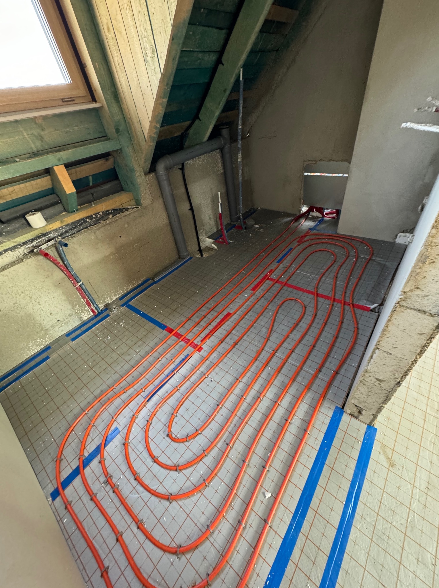 Pomarańczowe rury ogrzewania podłogowego ułożone na szarej macie w trakcie montażu w poddaszu, zabezpieczone niebieską taśmą, widoczne fragmenty instalacji hydraulicznej i okno dachowe.