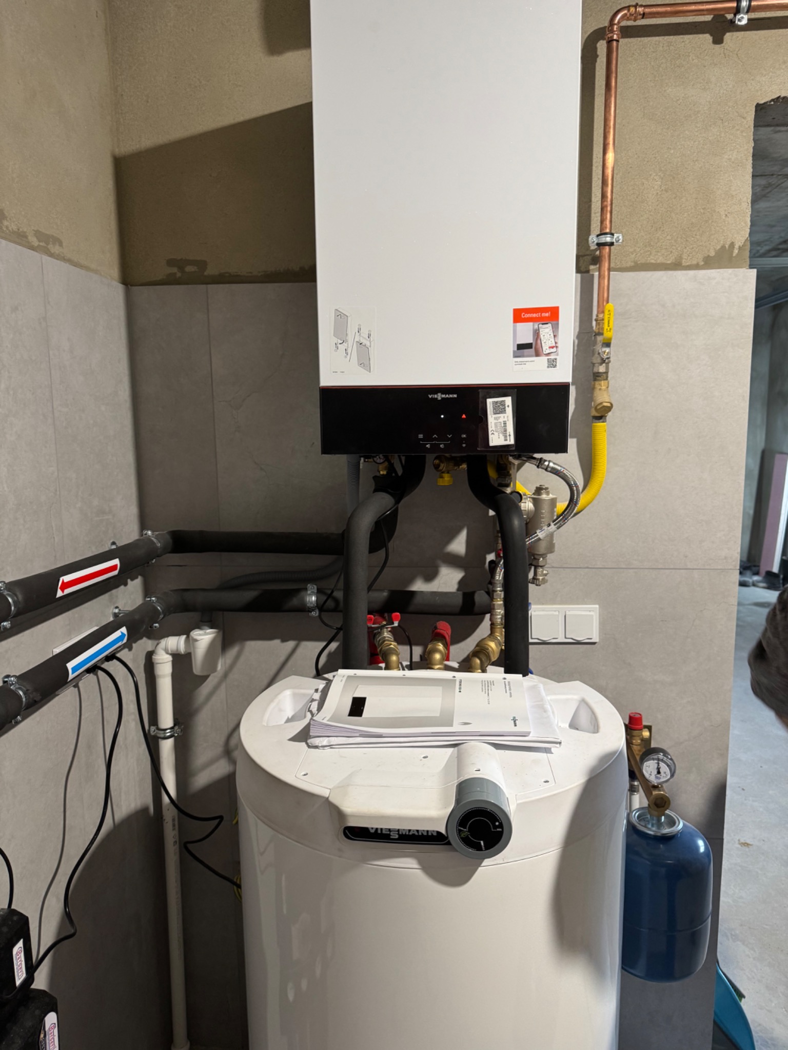 Nowoczesny piec gazowy Viessmann z zasobnikiem ciepłej wody, zamontowany w pomieszczeniu z szarymi płytkami, widoczne rury instalacji grzewczej i gazowej, oraz manometr na zasobniku.