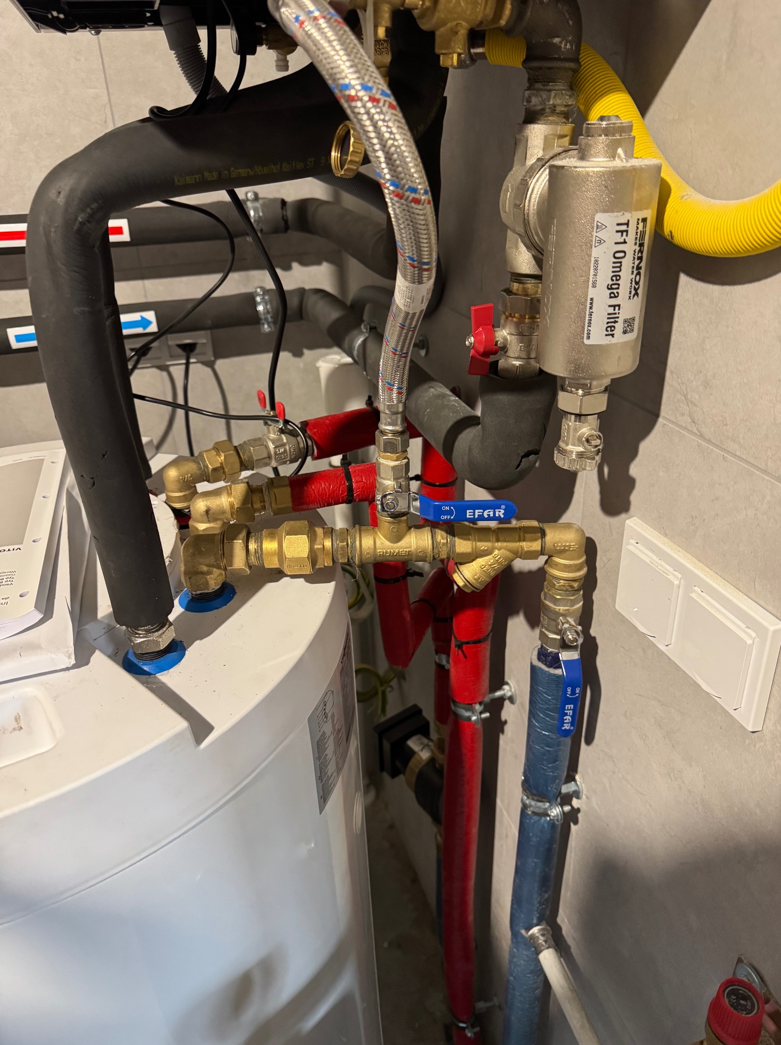 Instalacja grzewcza z widocznym filtrem Fernox TF1 Omega, rurami w izolacji czerwonej i niebieskiej, oraz elementami miedzianymi i zaworami. Widoczne również rury gazowe w żółtej osłonie karbowanej.