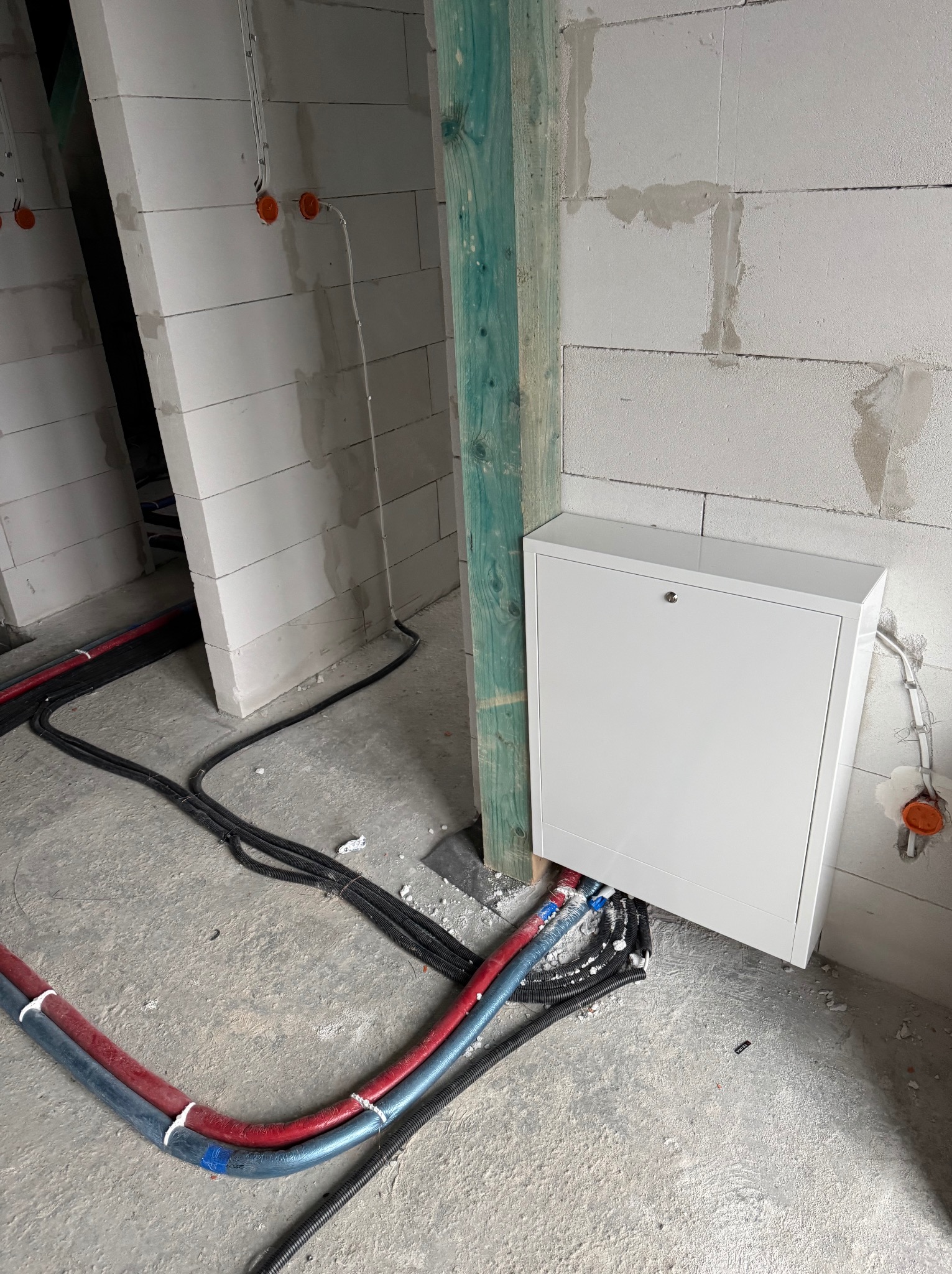 Biała skrzynka instalacyjna z zamkiem, zamontowana na surowej ścianie z betonu komórkowego, widoczne rury i peszle instalacyjne, drewniana belka konstrukcyjna.