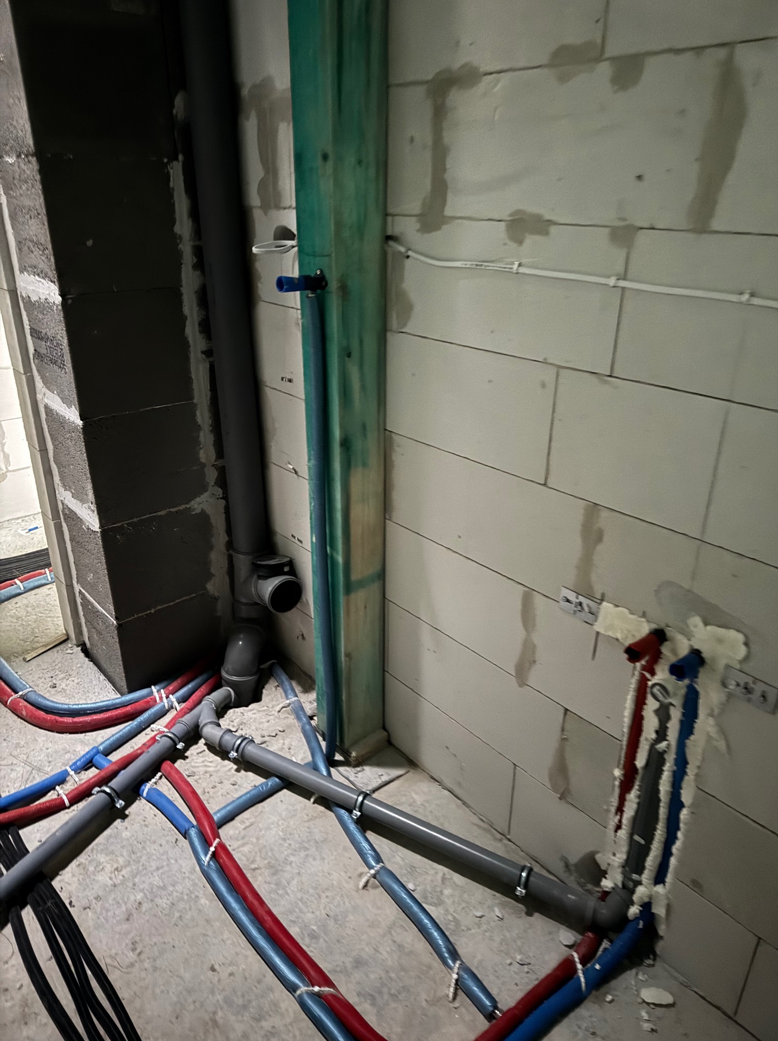Wnętrze w trakcie budowy z widocznymi rurami instalacji wodnej (czerwone i niebieskie) oraz kanalizacyjnymi (szare) na podłodze i w ścianie z betonu komórkowego, przymocowanymi do ściany za pomocą...