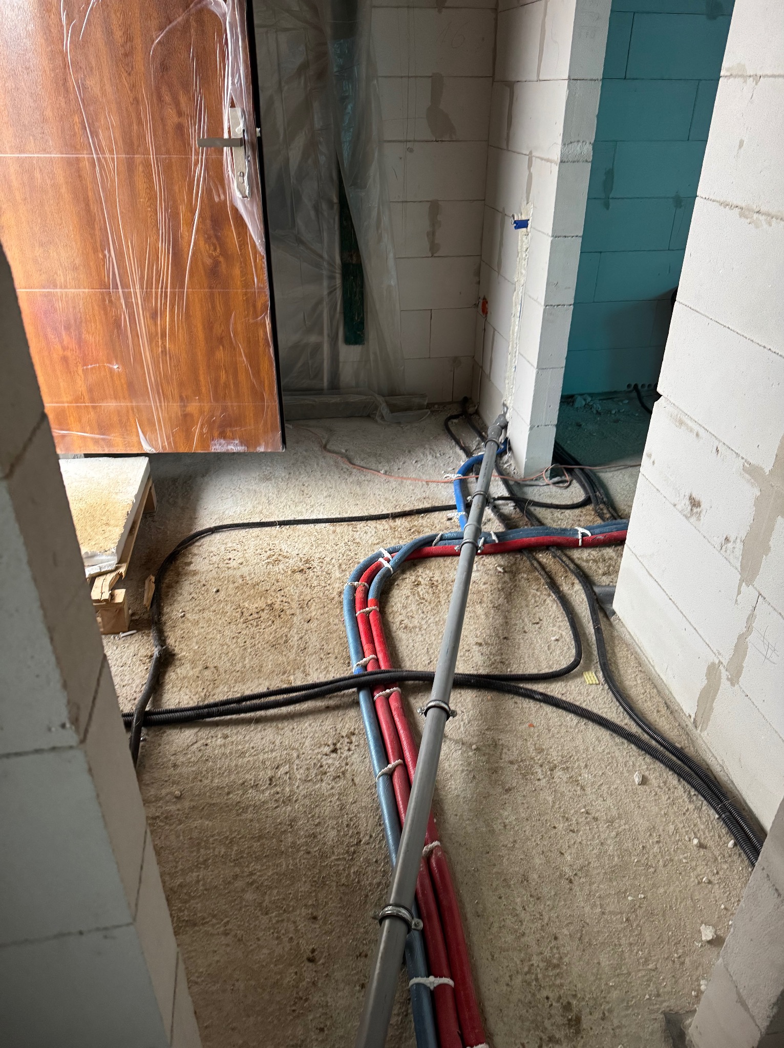 Wnętrze w trakcie budowy z widocznymi rurami instalacji grzewczej i hydraulicznej ułożonymi na surowej podłodze, ściany z białych bloczków, częściowo folia ochronna na drzwiach.