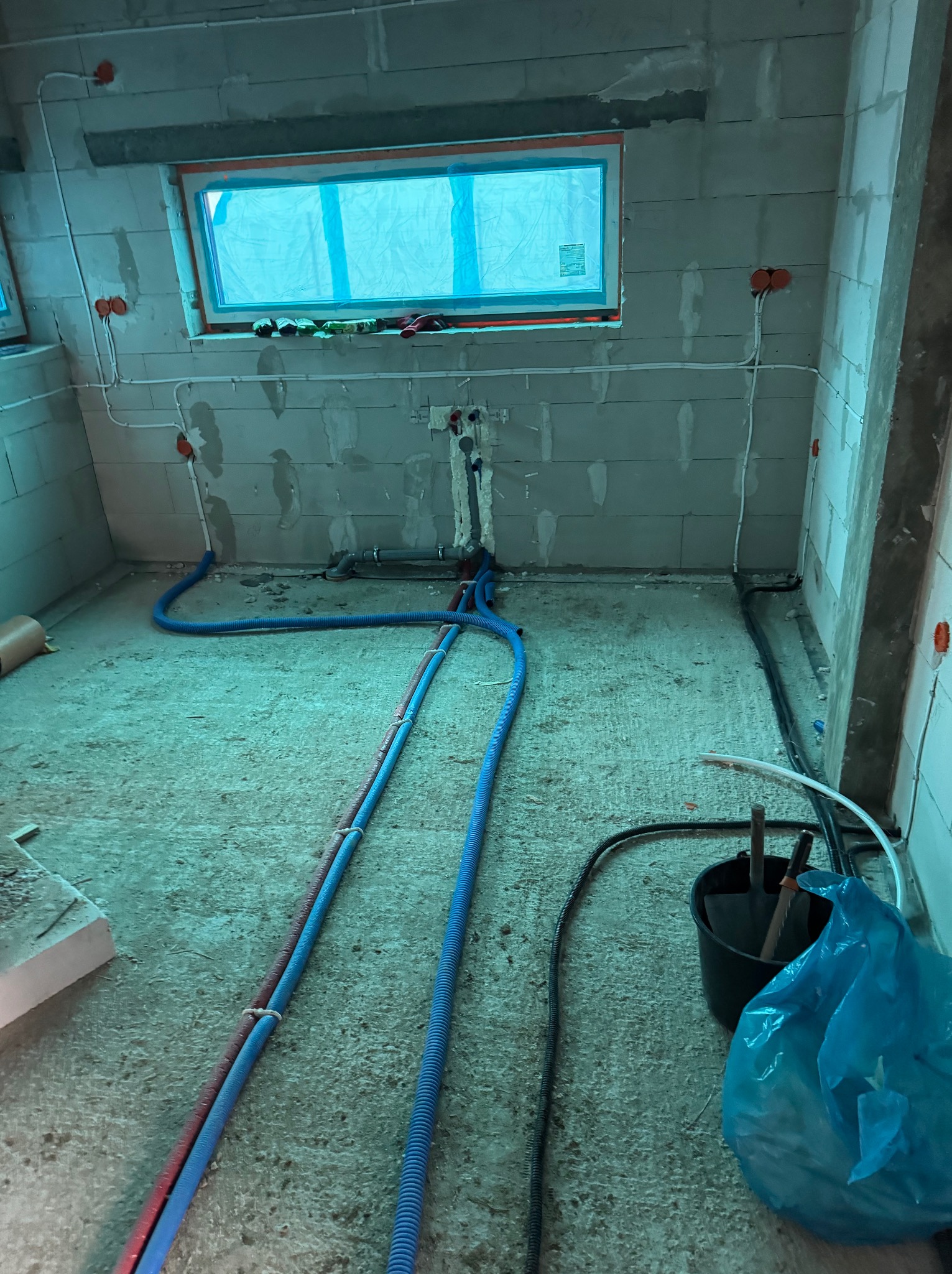 Surowy stan pomieszczenia w trakcie prac instalacyjnych: widoczne rury instalacji wodnej w niebieskiej karbowanej osłonie, poprowadzone po podłodze i ścianach z surowego betonu, puszki elektryczne...