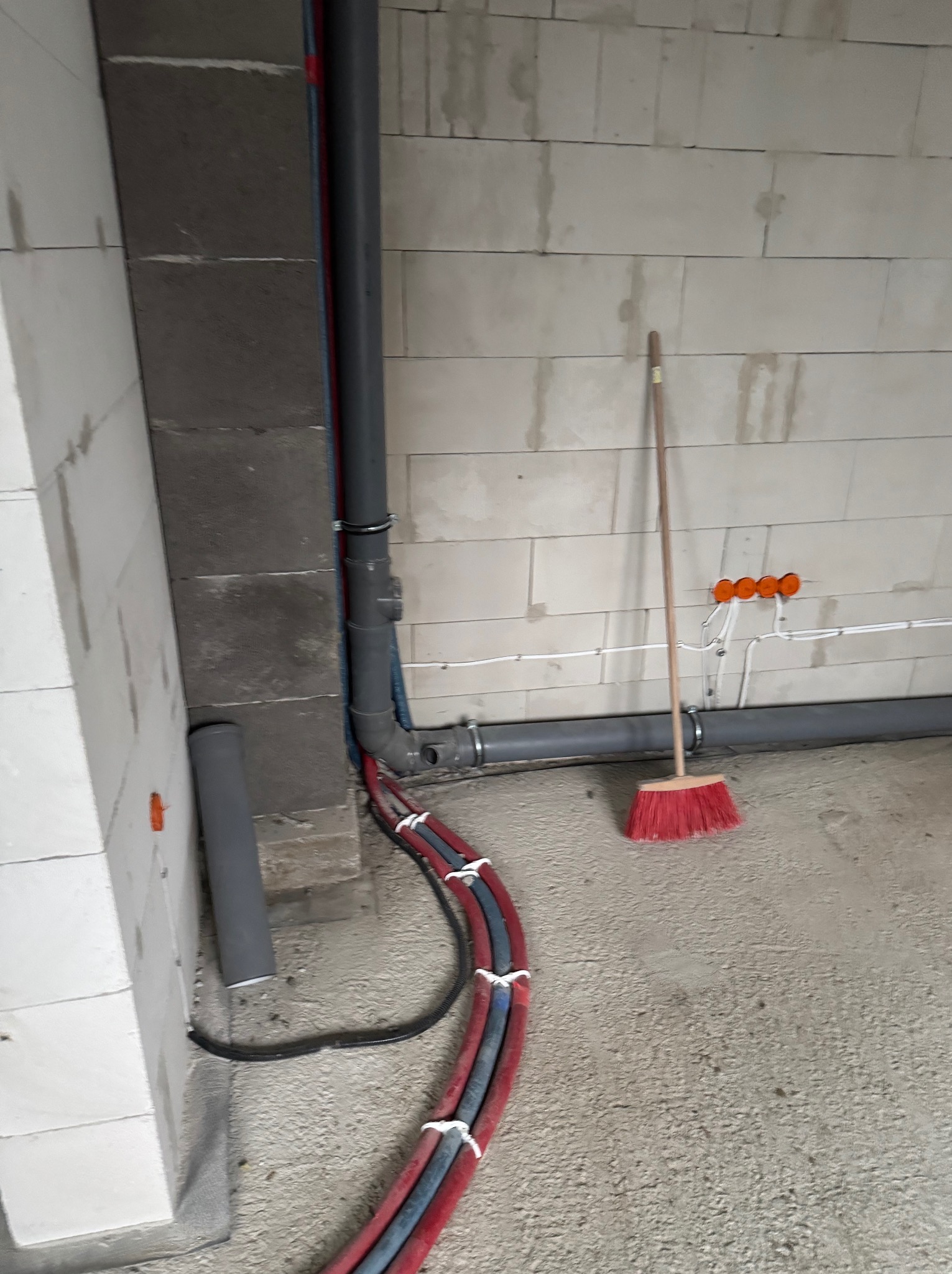 Wnętrze w trakcie prac instalacyjnych: widoczne rury grzewcze, kanalizacyjne, instalacja elektryczna oraz czerwona zmiotka oparta o ścianę z bloczków.
