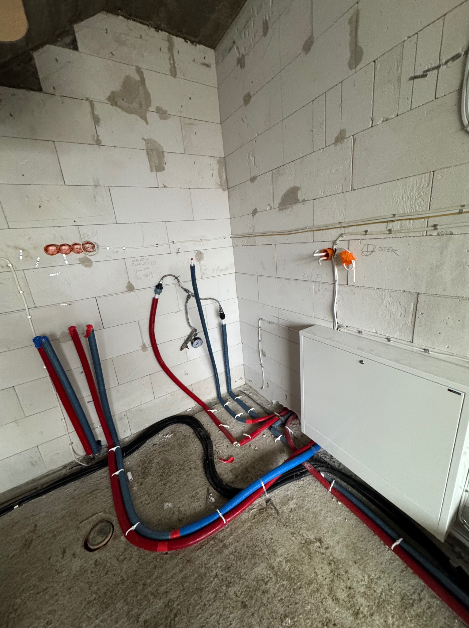 Surowe wnętrze w trakcie prac instalacyjnych: widoczne rury grzewcze i hydrauliczne w kolorach czerwonym i niebieskim, prowadzone po podłodze i ścianach z betonu komórkowego. Na ścianie widoczne...
