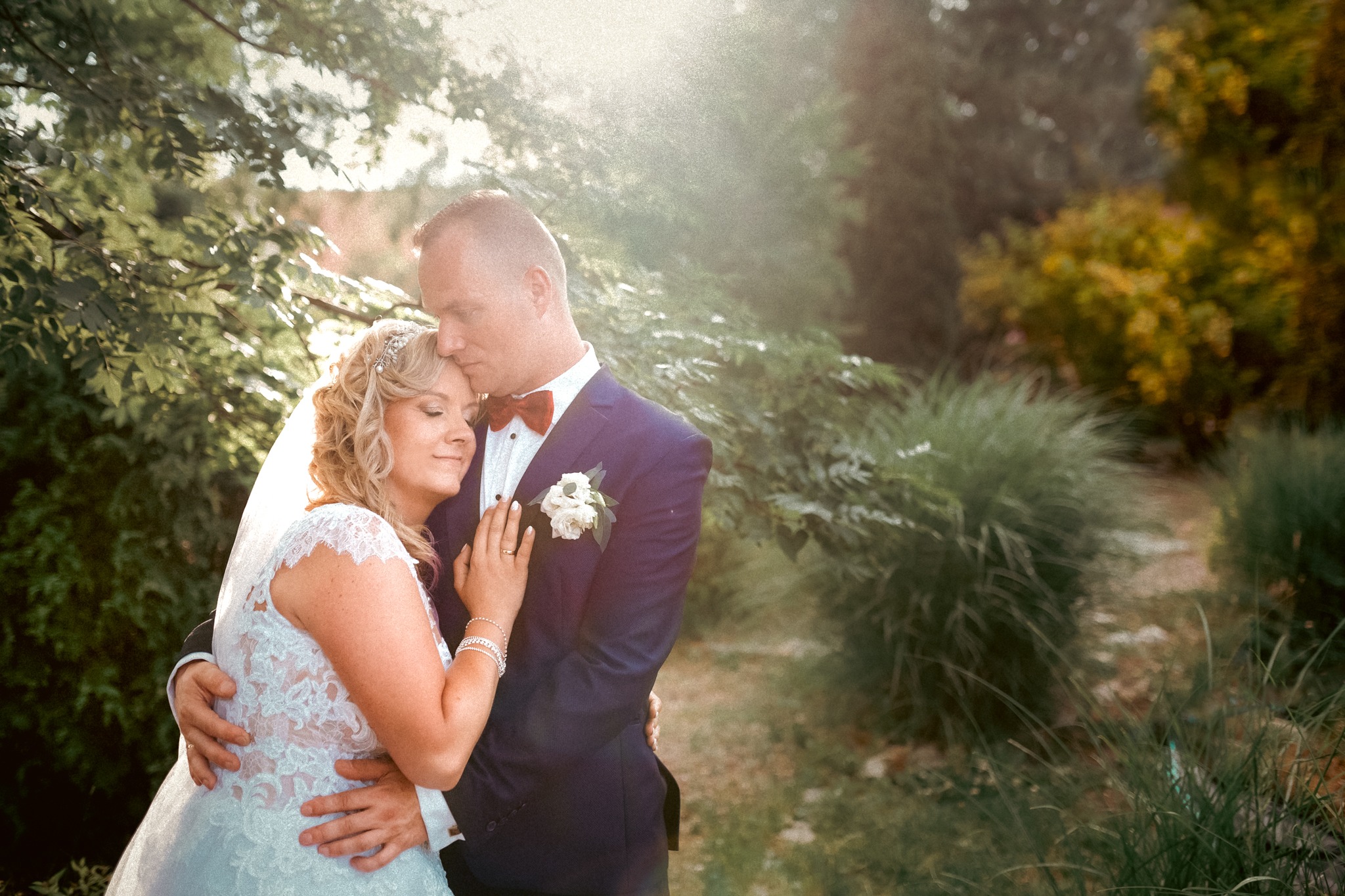 Para młoda przytulona w plenerze, panna młoda w koronkowej sukni i welonie, pan młody w granatowym garniturze z bordową muchą, ciepłe światło słoneczne.