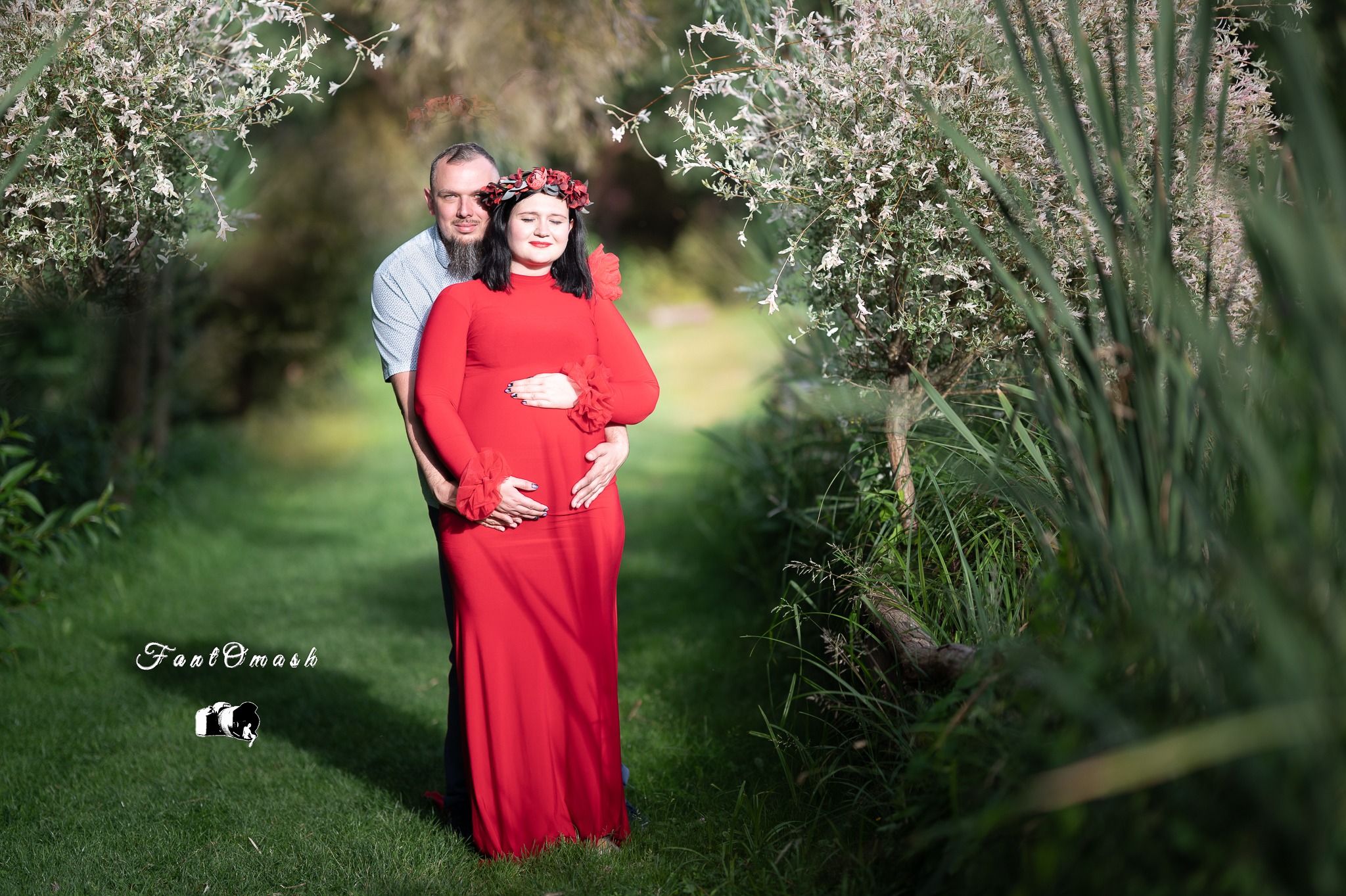 Kobieta w czerwonej sukni i wianku na głowie, obejmowana przez mężczyznę, pozuje na tle zieleni w plenerze.