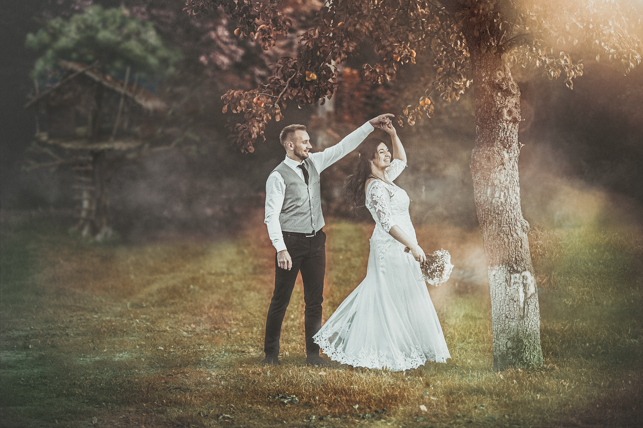 Para młoda tańczy pod drzewem z owocami w jesiennej scenerii, w tle widoczny domek na drzewie.