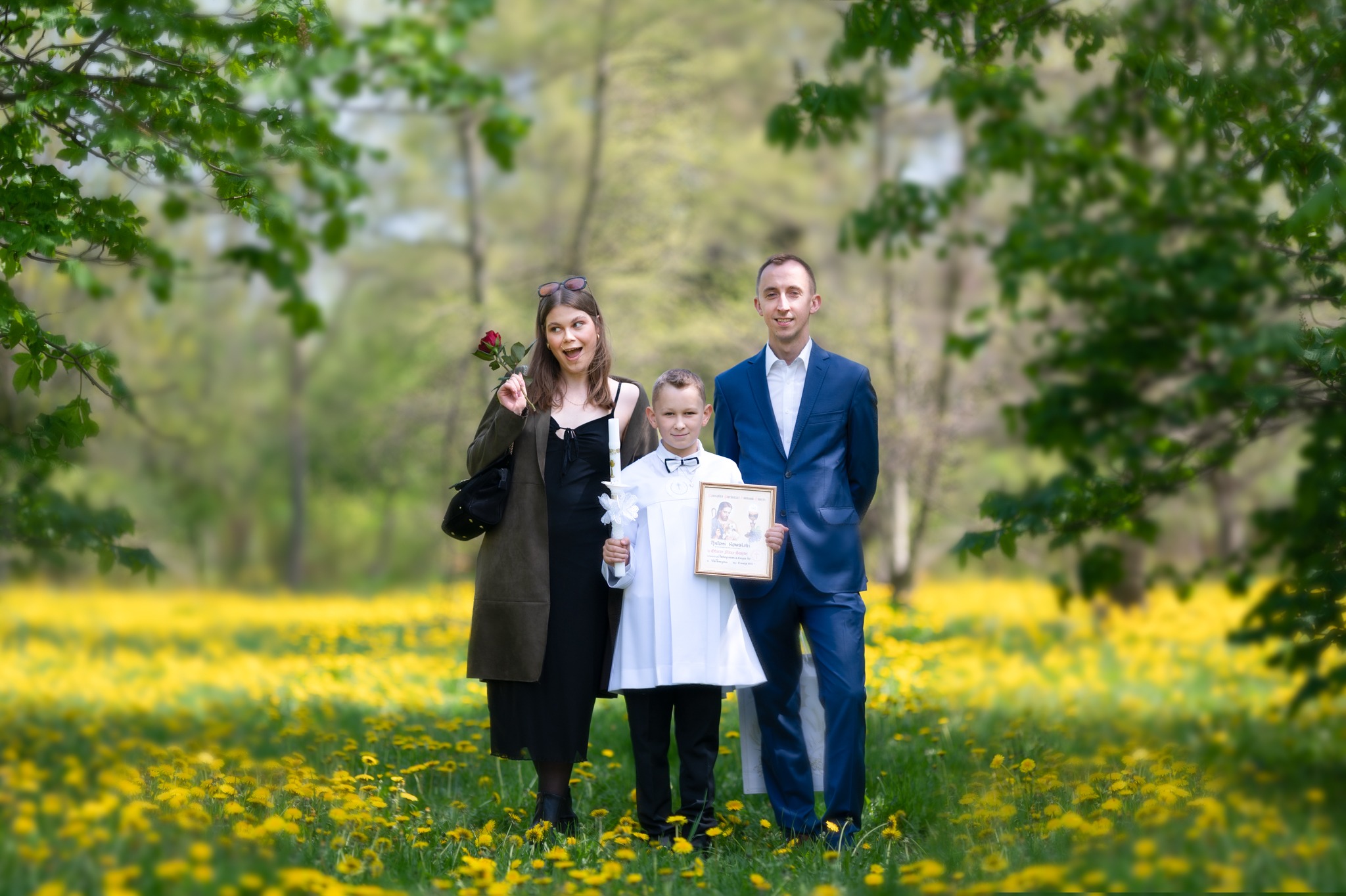 Rodzinne zdjęcie w plenerze z chłopcem w stroju komunijnym trzymającym obrazek, na tle łąki z kwitnącymi mleczami.
