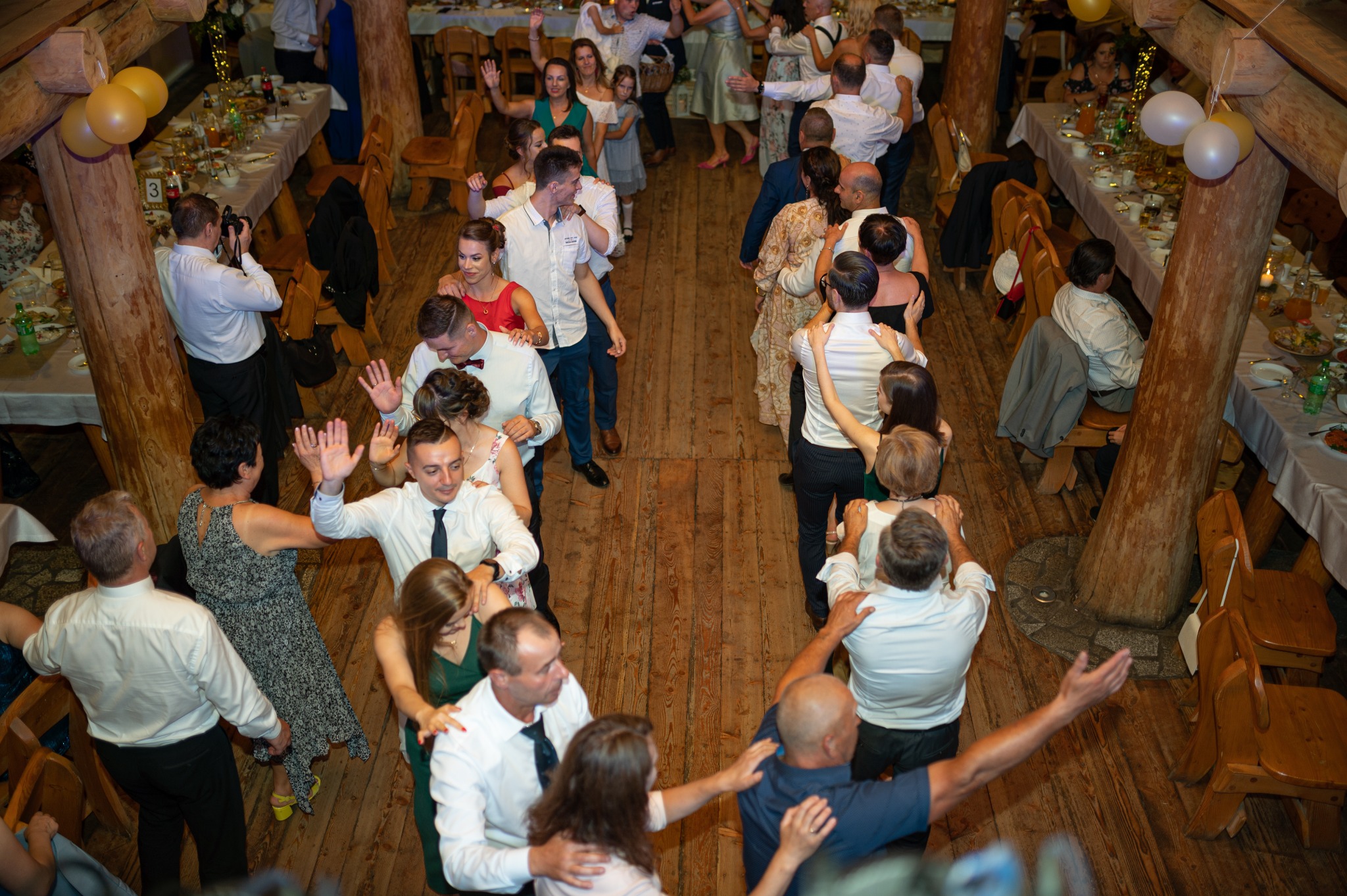 Widok z góry na salę weselną z drewnianymi elementami, gdzie goście tańczą między stołami, a jeden z gości robi zdjęcia aparatem.