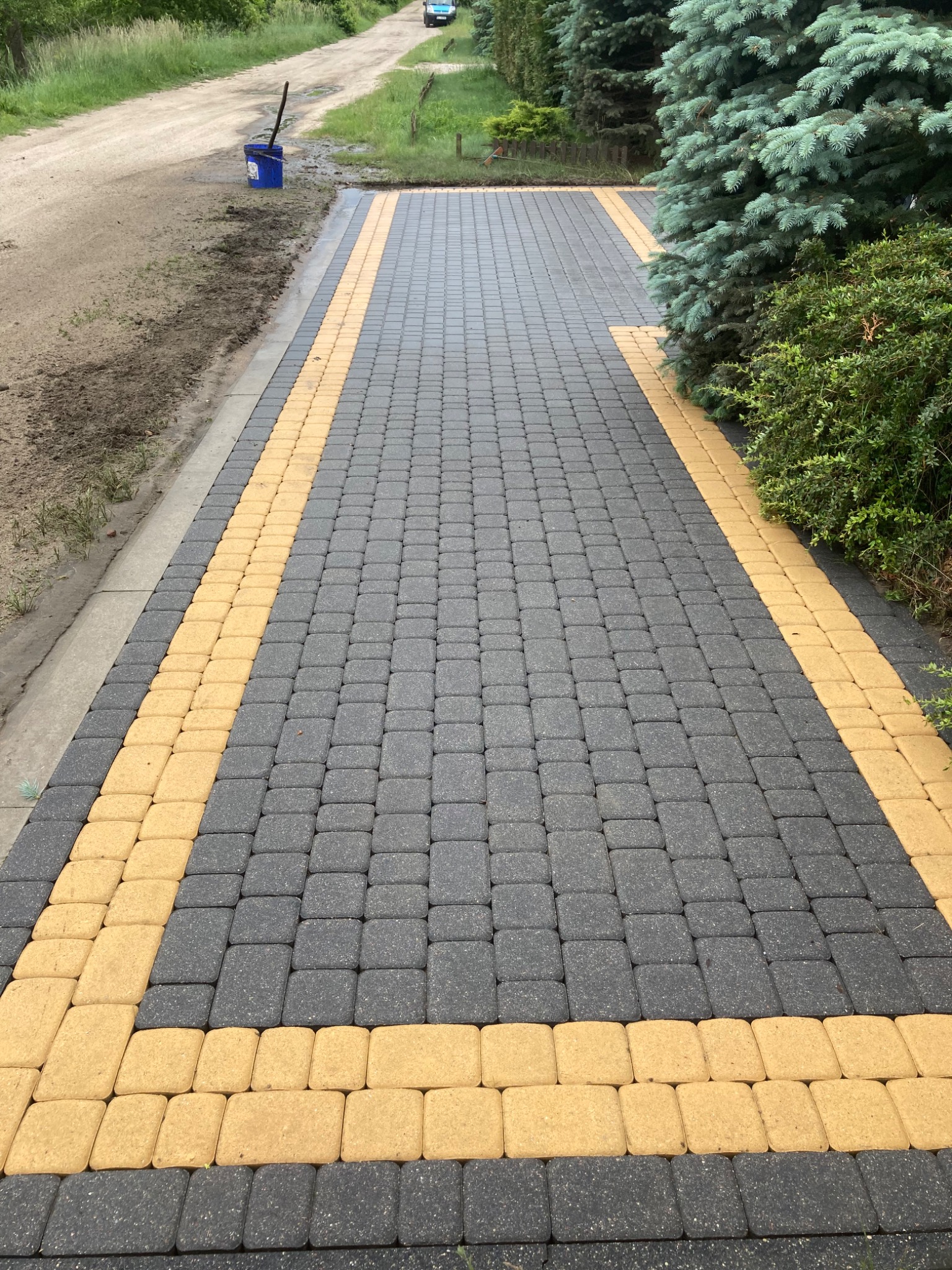 Ułożona kostka brukowa w dwóch kolorach: szarym i żółtym, tworząca wzór na podjeździe, widoczny niebieski pojemnik z narzędziami na uboczu.
