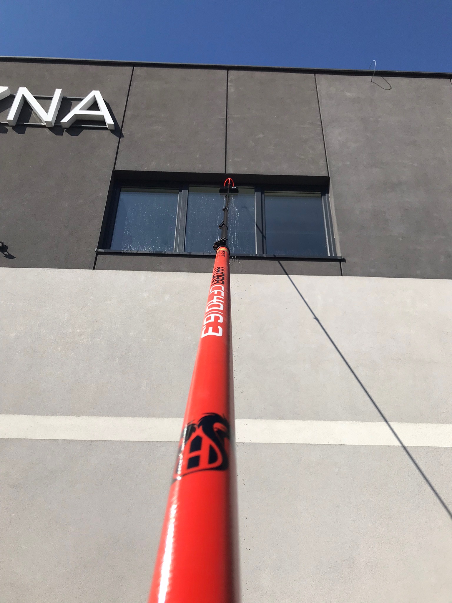 Czyszczenie okien na wysokości za pomocą teleskopowego kija z dyszą, widoczne krople spływającej wody na szybie, szara elewacja budynku z białym napisem.