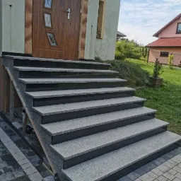 Zewnętrzne schody wejściowe z szarego kamienia z czarnymi obrzeżami, prowadzące do brązowych drzwi frontowych z dekoracyjnymi szybami, widoczne z boku na tle zielonego trawnika i domu.