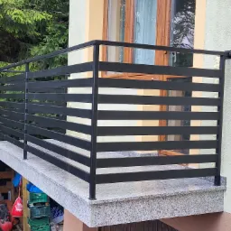 Balkon z balustradą z czarnych, poziomych profili i wykończeniem z jasnego, nakrapianego kamienia, widok z zewnątrz na tle zieleni.