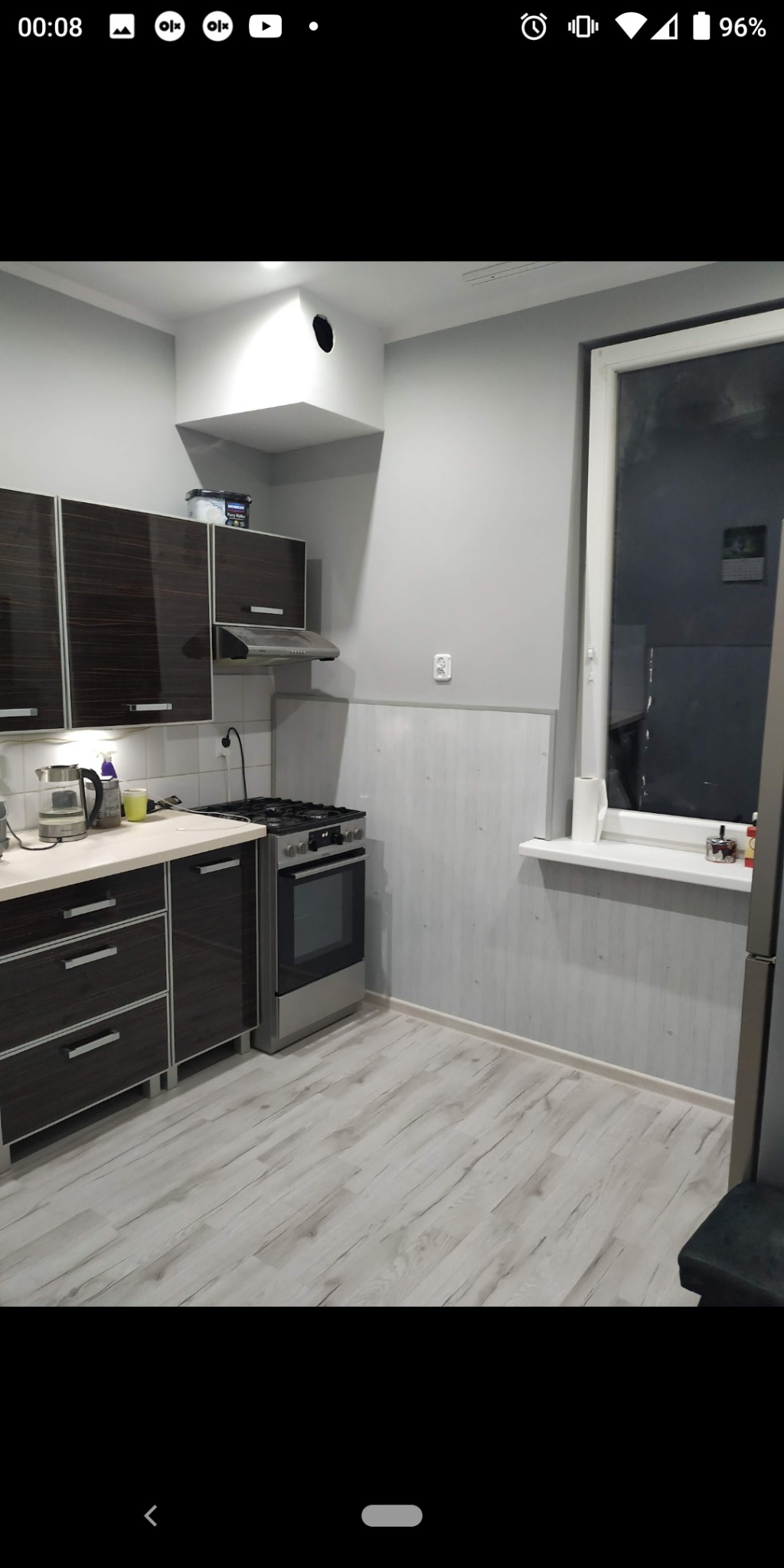 Kuchnia po remoncie: brązowe szafki z połyskiem, jasnoszare ściany, panele podłogowe imitujące drewno, widoczny fragment okna i kuchenki gazowej z piekarnikiem.