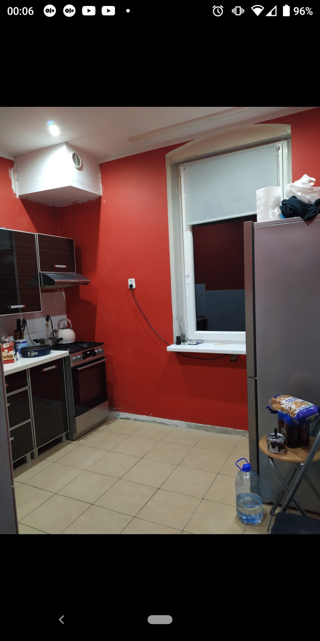 Kuchnia przed remontem: czerwona ściana z widocznymi śladami po pracach wykończeniowych, okno z roletą, meble kuchenne i sprzęty.