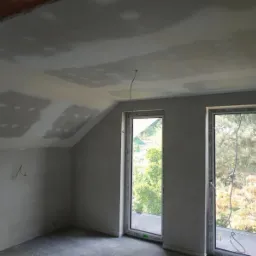 Pokój na poddaszu w trakcie prac wykończeniowych: ściany i sufit po wstępnym szpachlowaniu, widoczne dwa okna dachowe z widokiem na drzewa.