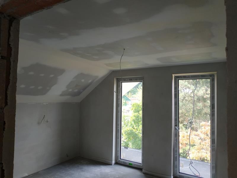 Pokój na poddaszu w trakcie prac wykończeniowych: ściany i sufit po wstępnym szpachlowaniu, widoczne dwa okna dachowe z widokiem na drzewa.