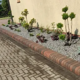 Ogród przydomowy z ozdobnymi krzewami i roślinami, wysypany szarym kamieniem, oddzielony od chodnika ceglanym obrzeżem.