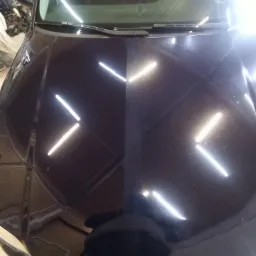 Ciemny lakierowany fragment karoserii samochodu, z widocznym odbiciem lamp, po polerowaniu, z wyraźnym podziałem na część wypolerowaną i niewypolerowaną.