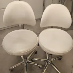 Dwa białe, skórzane krzesła obrotowe na metalowych podstawach z kółkami, ustawione obok siebie na szarej podłodze. Widoczne ślady użytkowania na siedziskach.