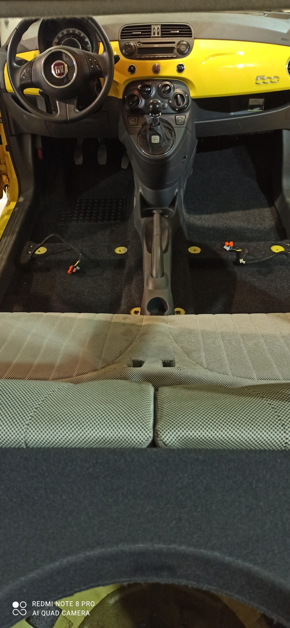 Wnętrze żółtego Fiata 500 z częściowo zdemontowaną tapicerką podłogową, widoczne kable i elementy montażowe.