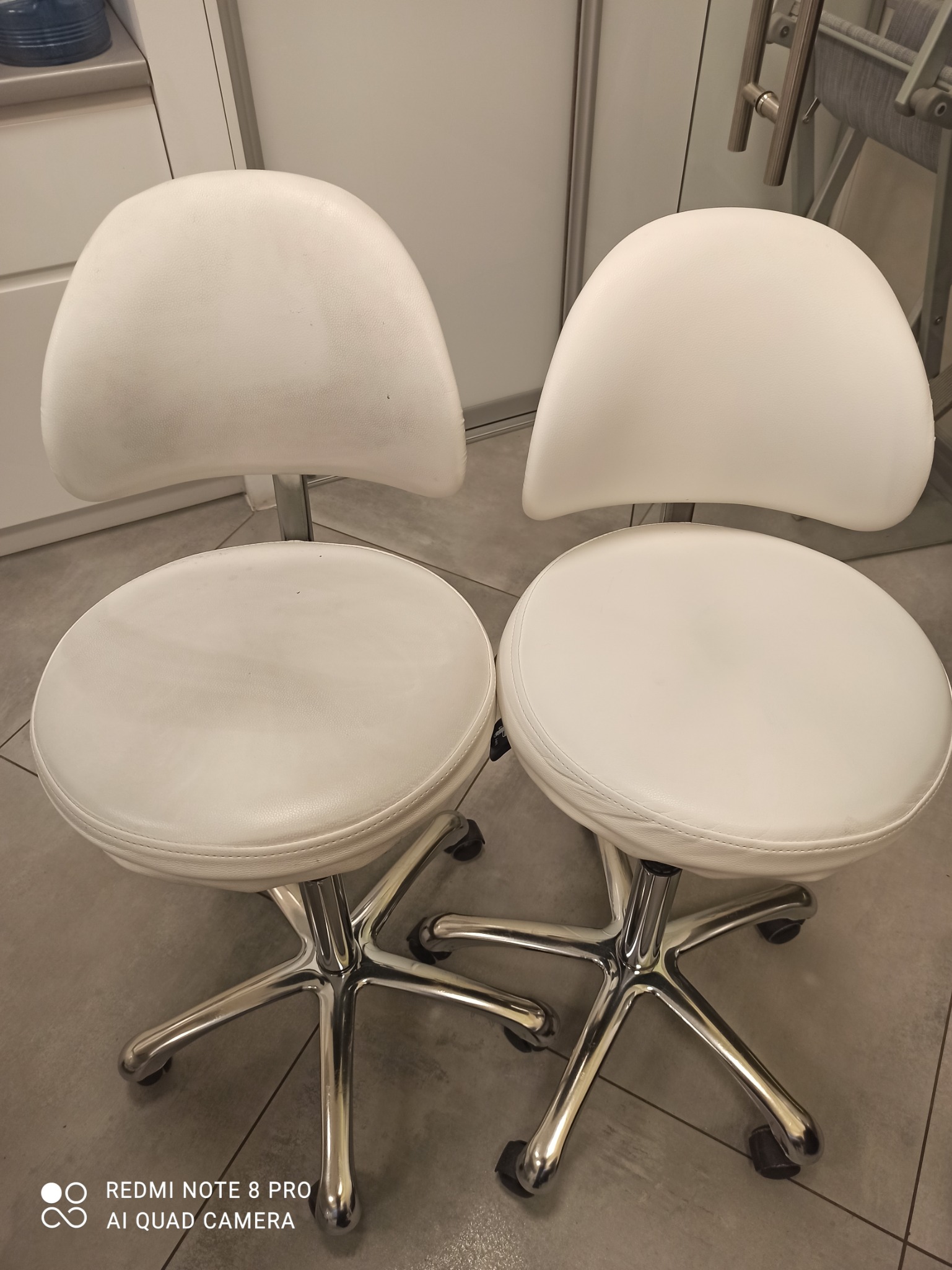 Dwa białe, skórzane krzesła obrotowe na metalowych podstawach z kółkami, ustawione obok siebie na szarej podłodze. Widoczne ślady użytkowania na siedziskach.